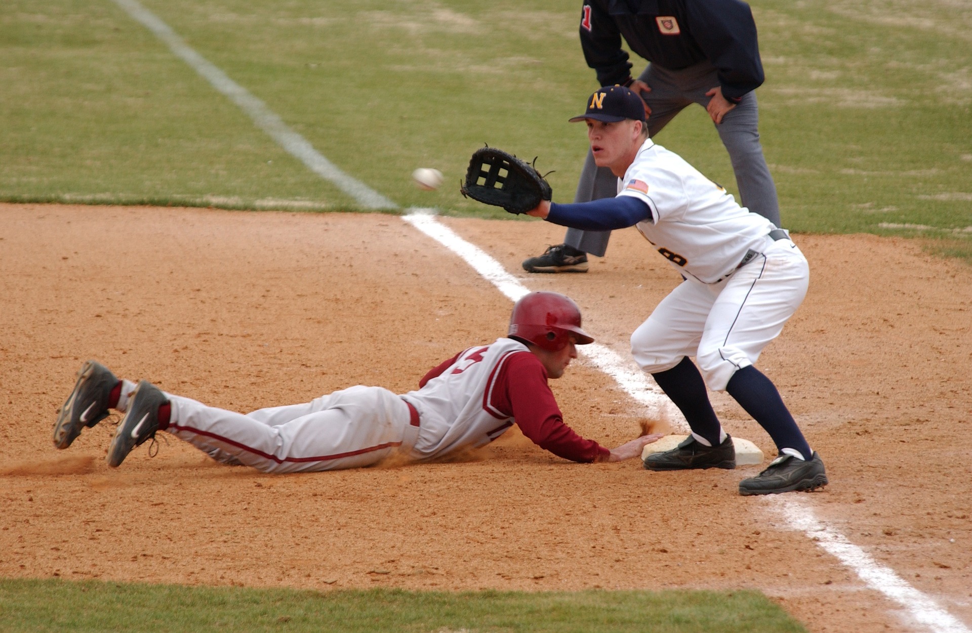 Runner sliding in head first to first base where the first basemen catcher is just about to catch the ball: Types of Baseball Gloves [MindfuseBaseball.com]