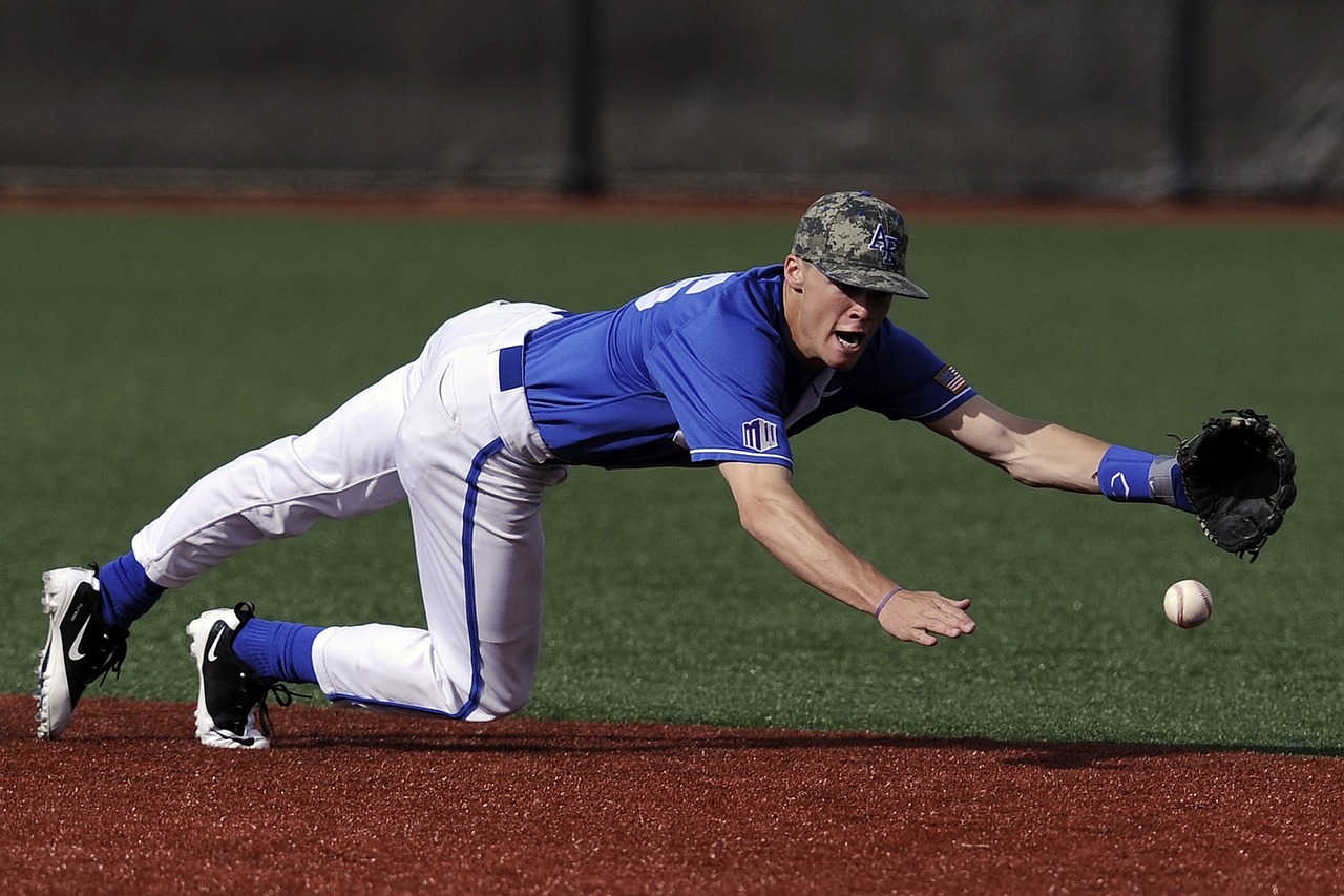 Baseball infielder diving to catch the ball with his left hand. He is wearing a blu top and white bottoms with a green hat on: Types of Baseball Gloves [MindfuseBaseball.com]